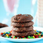 A stack of rich, fudgy brownie cookies studded with mini M&Ms, sitting on a parchment-lined wire rack with colorful candies scattered around.