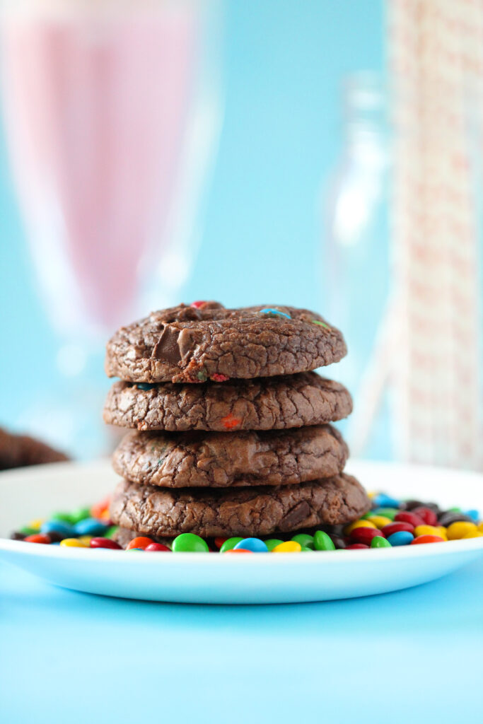 A stack of rich, fudgy brownie cookies studded with mini M&Ms, sitting on a parchment-lined wire rack with colorful candies scattered around.