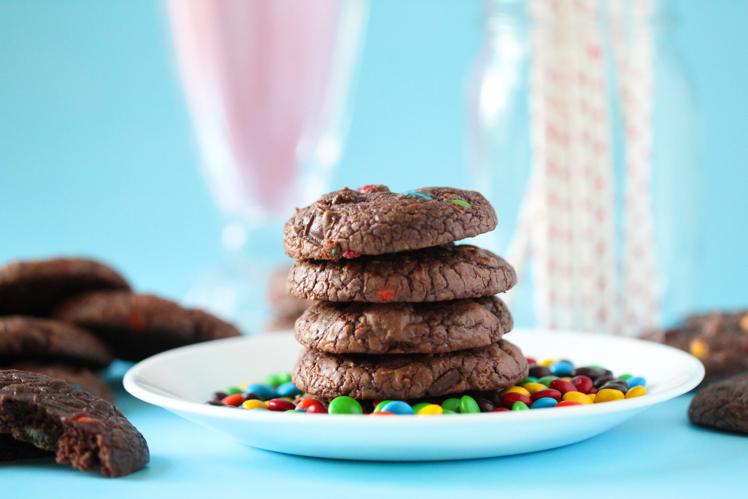 A stack of rich, fudgy brownie cookies studded with mini M&Ms, sitting on a parchment-lined wire rack with colorful candies scattered around.