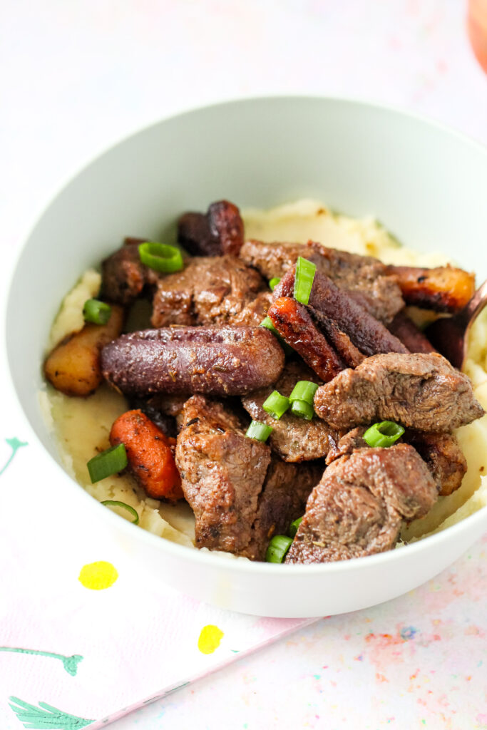 A bowl of garlic butter beef bites on top of mashed potatoes with carrots.
