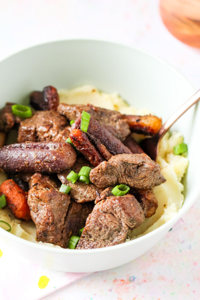 A bowl of garlic butter beef bites on top of mashed potatoes with carrots.