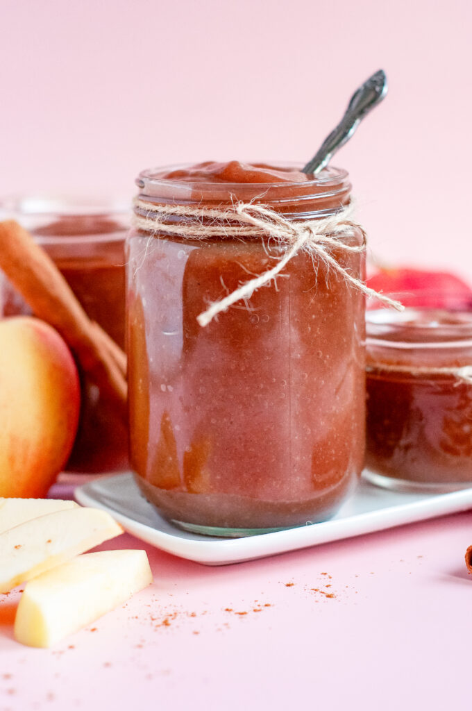 Jar of homemade caramel apple butter tied with twine, surrounded by fresh apples, cinnamon sticks, and apple slices on a pink background — a cozy fall spread perfect for serving warm with flaky Grands biscuits.