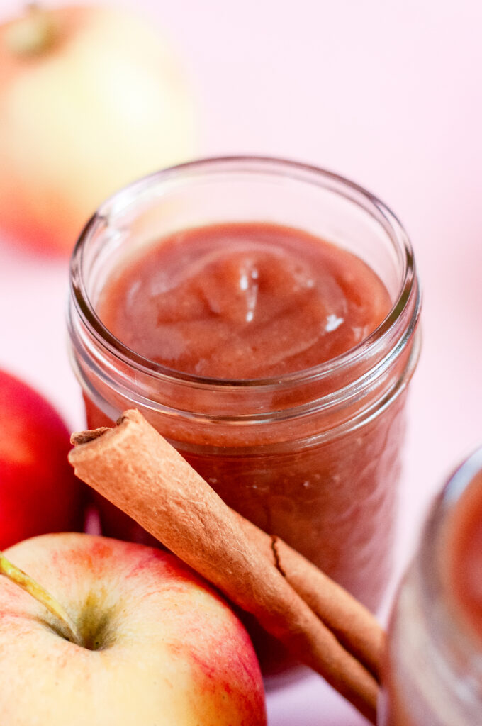 Jar of homemade caramel apple butter tied with twine, surrounded by fresh apples, cinnamon sticks, and apple slices on a pink background — a cozy fall spread perfect for serving warm with flaky Grands biscuits.