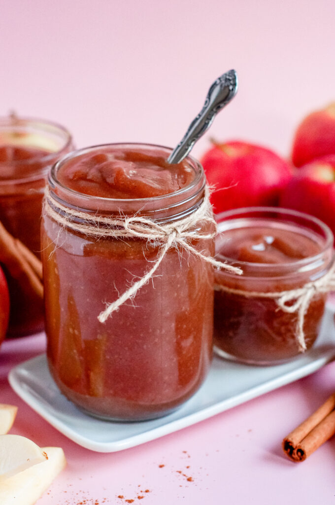 Jar of homemade caramel apple butter tied with twine, surrounded by fresh apples, cinnamon sticks, and apple slices on a pink background — a cozy fall spread perfect for serving warm with flaky Grands biscuits.