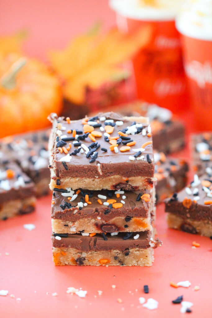 Chocolate-frosted cookie bars topped with orange, white, and black Halloween sprinkles shaped like ghosts, bats, and hearts, arranged on a coral-colored surface.