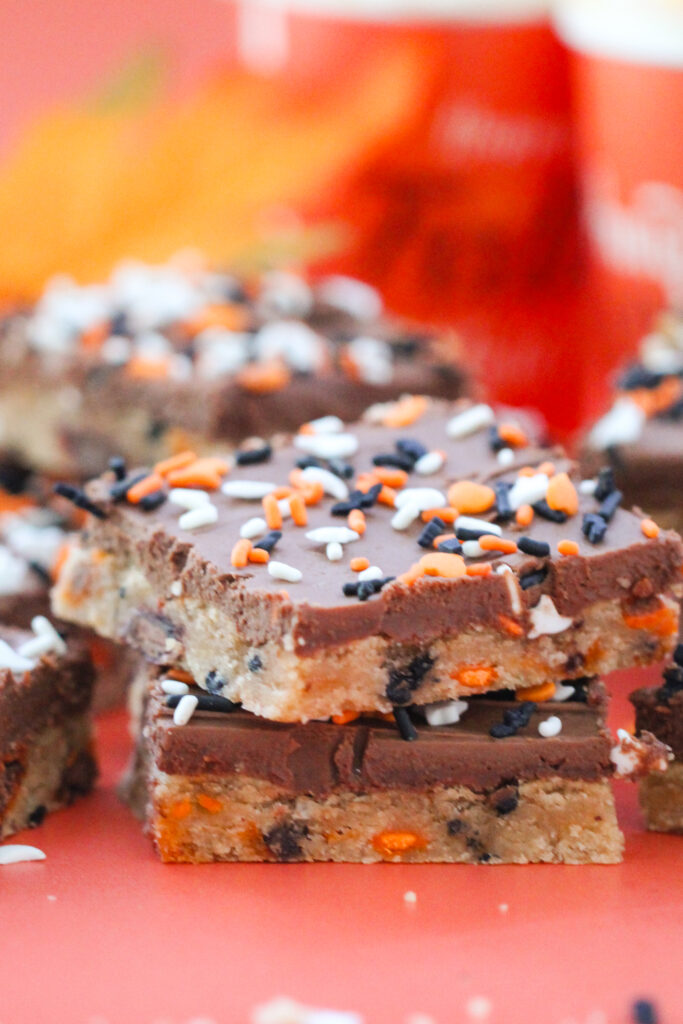 Chocolate-frosted cookie bars topped with orange, white, and black Halloween sprinkles shaped like ghosts, bats, and hearts, arranged on a coral-colored surface.