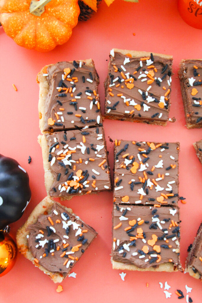 Chocolate-frosted cookie bars topped with orange, white, and black Halloween sprinkles shaped like ghosts, bats, and hearts, arranged on a coral-colored surface.