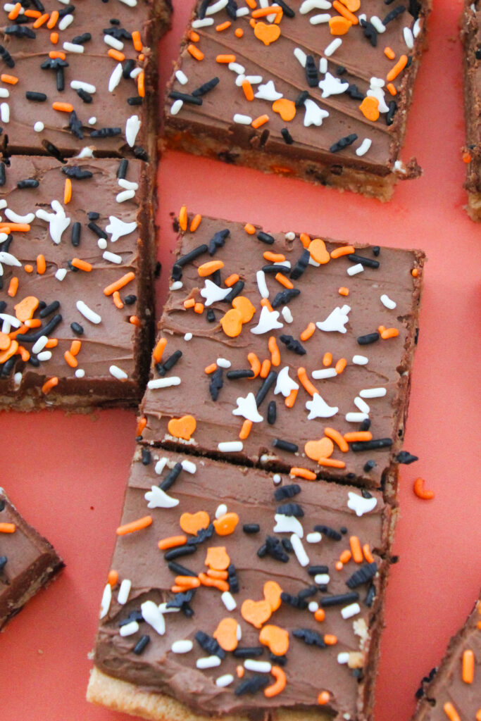 Chocolate-frosted cookie bars topped with orange, white, and black Halloween sprinkles shaped like ghosts, bats, and hearts, arranged on a coral-colored surface.