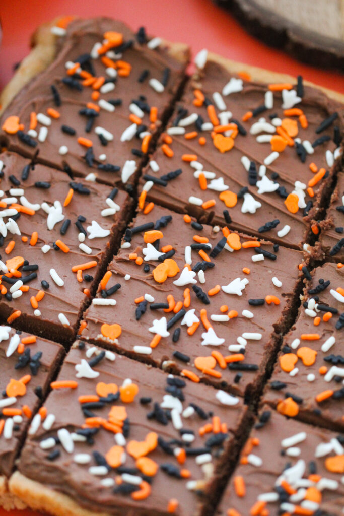 Chocolate-frosted cookie bars topped with orange, white, and black Halloween sprinkles shaped like ghosts, bats, and hearts, arranged on a coral-colored surface.
