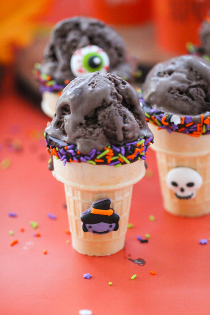 Halloween-themed ice cream cones filled with scoops of dark chocolate ice cream, decorated with orange, purple, and green sprinkles, candy eyeballs, and tiny bats, witch, and skeleton decorations, set against a bright orange background with pumpkins and festive treats.
