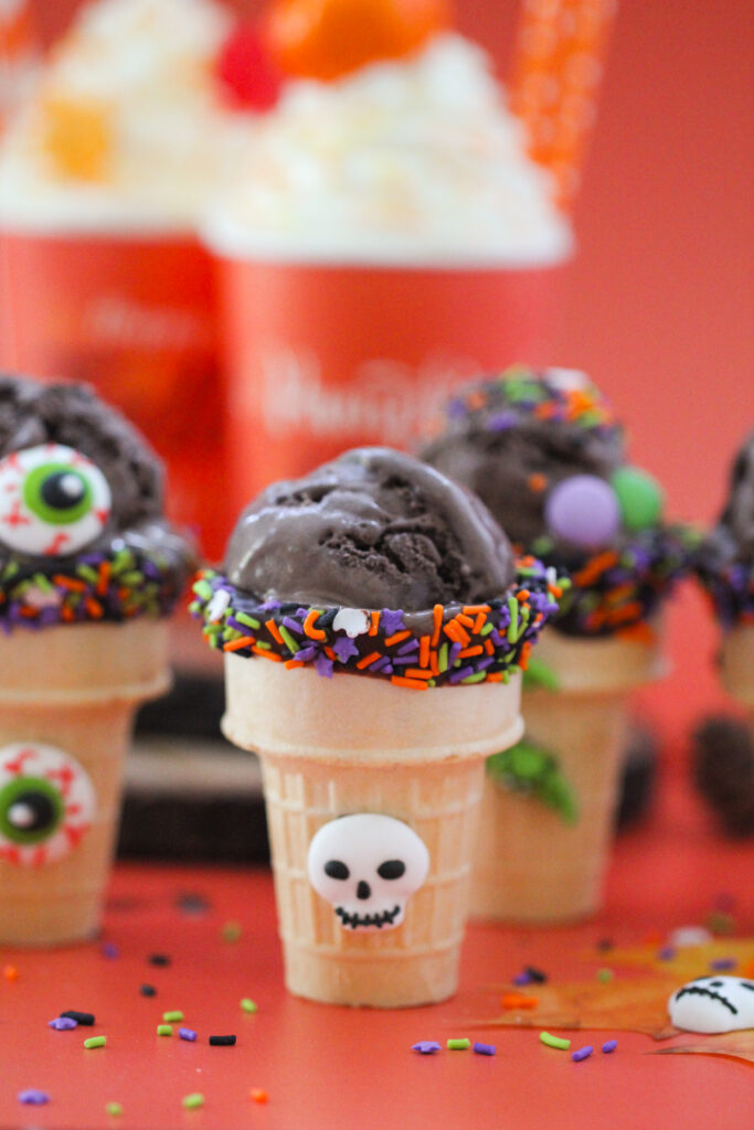 Halloween-themed ice cream cones filled with scoops of dark chocolate ice cream, decorated with orange, purple, and green sprinkles, candy eyeballs, and tiny bat, witch, and skeleton decorations, set against a bright orange background with pumpkins and festive treats.