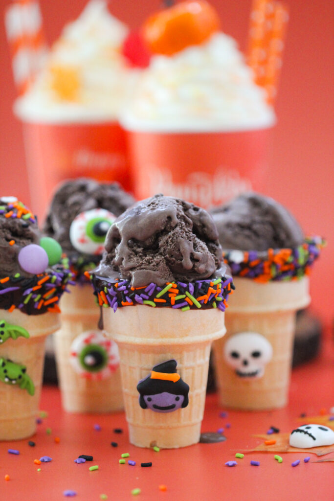 Halloween-themed ice cream cones filled with scoops of dark chocolate ice cream, decorated with orange, purple, and green sprinkles, candy eyeballs, and tiny bats, witch, and skeleton decorations, set against a bright orange background with pumpkins and festive treats.