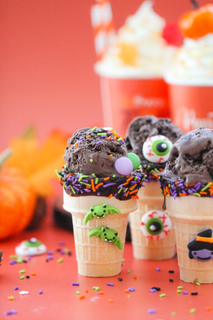 Halloween-themed ice cream cones filled with scoops of dark chocolate ice cream, decorated with orange, purple, and green sprinkles, candy eyeballs, and tiny bats, witch, and skeleton decorations, set against a bright orange background with pumpkins and festive treats.
